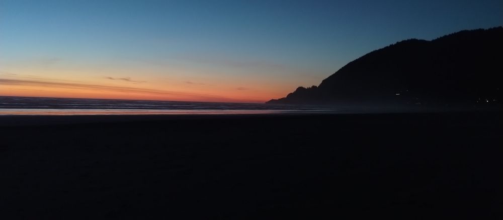 Atmospheric photo of a late sunset on the beach at Manzanita, OR, USA. The sky is a moody late sunset, oranges at the horizon line, lightening to white and then ombre from white to deep navy blue at the top of the picture. On the right side, the edge of a small mountain slopes into the waves visible in the middle distance. An expanse of firm sand fills the frame about midway up.