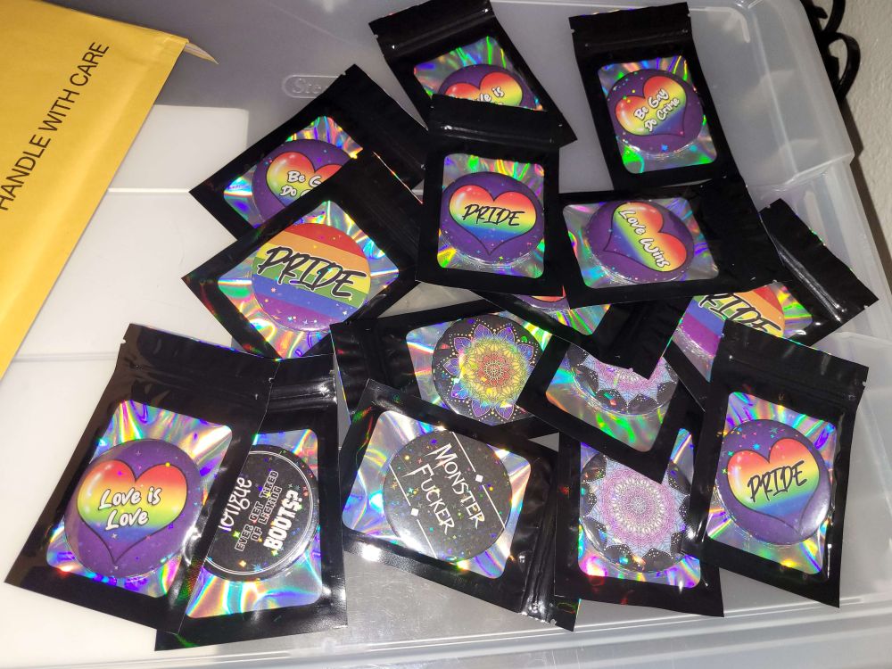 A decent amount of Pride themed buttons in holographic and black bags. Some are hearts saying love is love, pride, be gay do crime, love wins. Others are trans and pride mandalas. Two are goth sayings of Monster Fucker and Does your tongue ever get tired of licking boots?