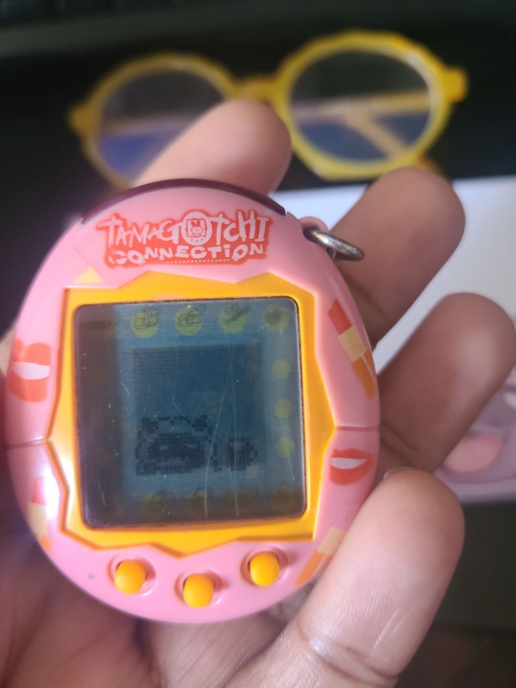 An image of me holding a pink tamagotchi connection that's shaped like an egg and has yellow buttons. There's a small screen inside it showing my tamagotchi ( a tiny sheep) brushing their teeth with a little mug to the right of them.