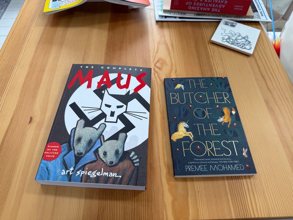 Two books on a wooden table. “Maus” by Art Spiegelman and “The Butcher of the Forest” by Premee Mohamed