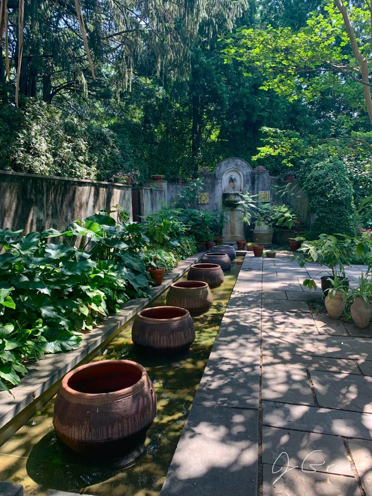 A photograph of an enclosed portion of Chanticleer Gardens. There are large clay pots in a long narrow pool of water. 