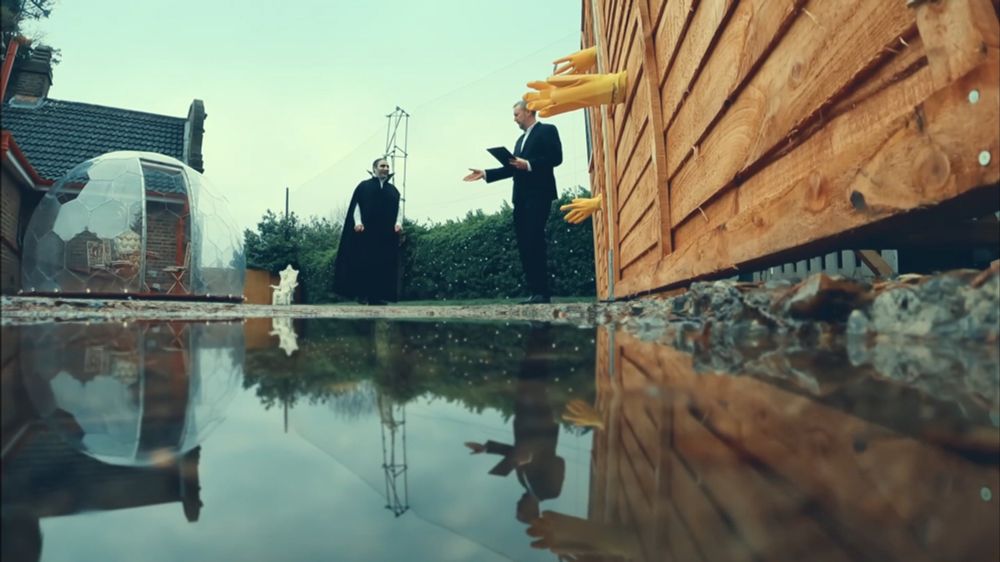 Screen grab of Taskmaster where Nick Mohammed is walking towards Alex Horne. Nick is dressed like Dracula. Alex is standing next to a wooden wall with yellow gloves sticking out. There's a big puddle of water in the frame, and we can see Alex's reflection but NOT Nick's, because Vampires don't have reflections.