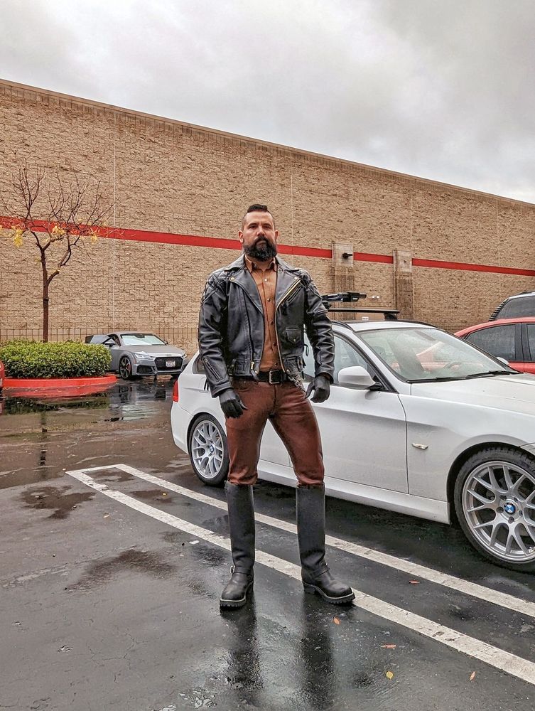 Jorge, a Latino man with a beard, standing in a parking lot in front of a white BMW wagon. He's wearing brown leather pants, a brown leather shirt, black tall leather Engineer boots, a heavy black leather jacket and black leather gloves. The pavement is wet and the sky is overcast.
