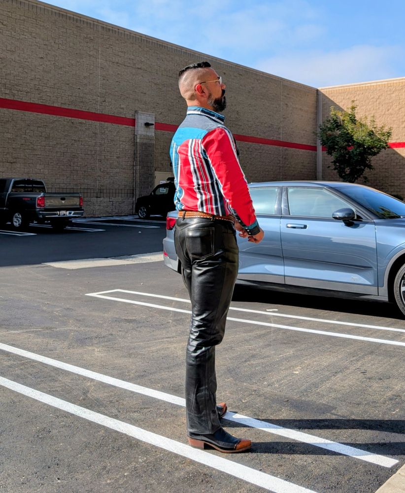 Jorge standing in a Costco parking lot. He's wearing black leather jeans, black cowboy boots with brown wingtips, a brown belt with a western buckle and a western snap long sleeve shirt with red, green, white, light blue and dark blue vertical stripes. 

He has a fresh haircut and beard trim. He's facing away from camera so the backside of his outfit can be seen. 