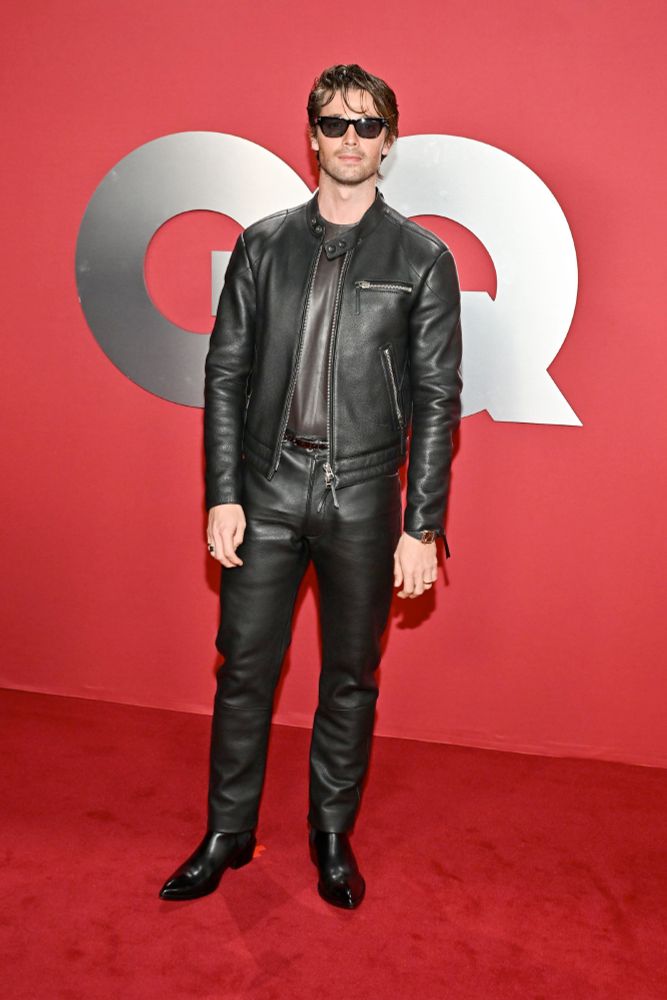 Patrick Schwarzenegger at a GQ red carpet. He's wearing leather pants, a leather jacket and a leather shirt, with black cowboy boots and sunglasses.