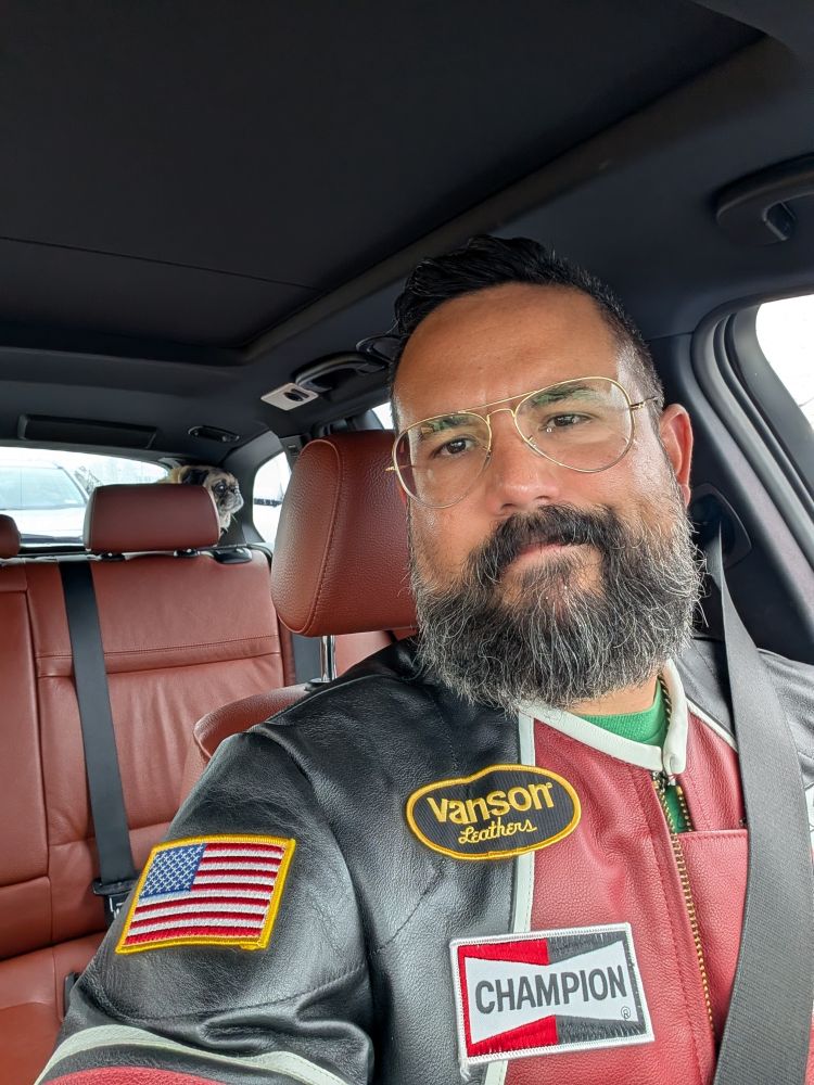 Selfie of Jorge in his car. He's wearing a red and dark blue leather jacket. Behind him, the back of his wagon shows Fede the fawn pug standing up looking out the rear side window. 