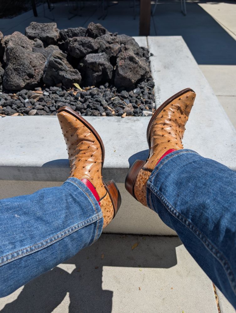 Tan colored ostrich cowboy boots propped up on a concrete fire pit. A little bit of a red leather shaft is peeking from under the blue jeans that cover them
