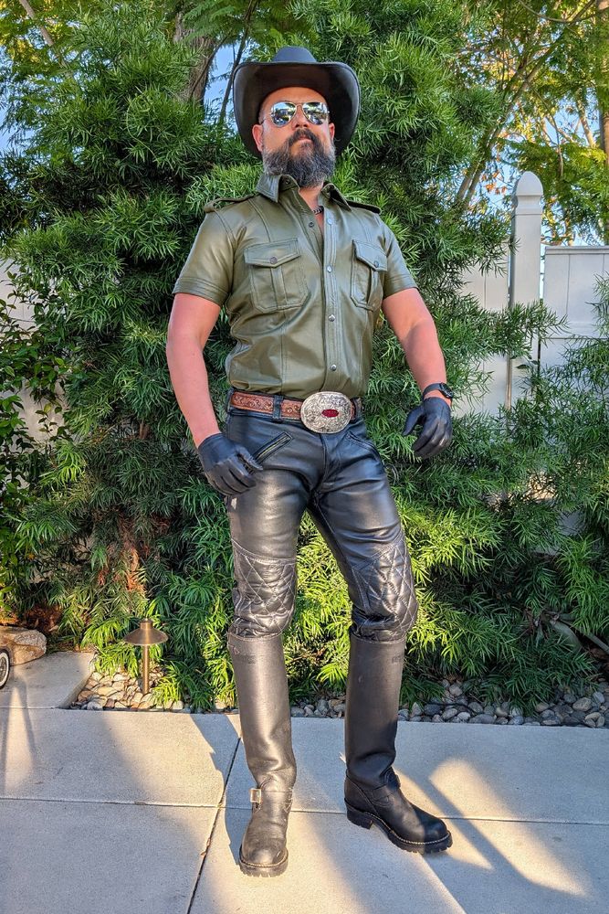 Jorge standing in a backyard wearing a short sleeve green leather shirt, black leather padded breeches, tall black leather Wesco engineer boots, a big silver belt buckle, a black leather cowboy hat, black leather short gloves and sunglasses.