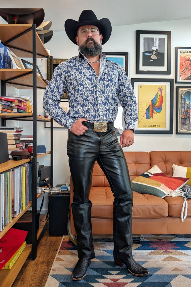 Jorge standing in front of a couch and a wall with artwork. 

He's wearing shiny black leather pants that thug his legs nicely. He has a white and blue Paisley patterned western snap shirt, a black cowboy hat, dark blue ostrich cowboy boots and a big silver and gold western buckle