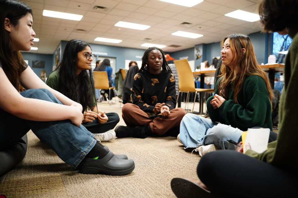 A group of four students are sitting on the floor in a circle talking about research. 
