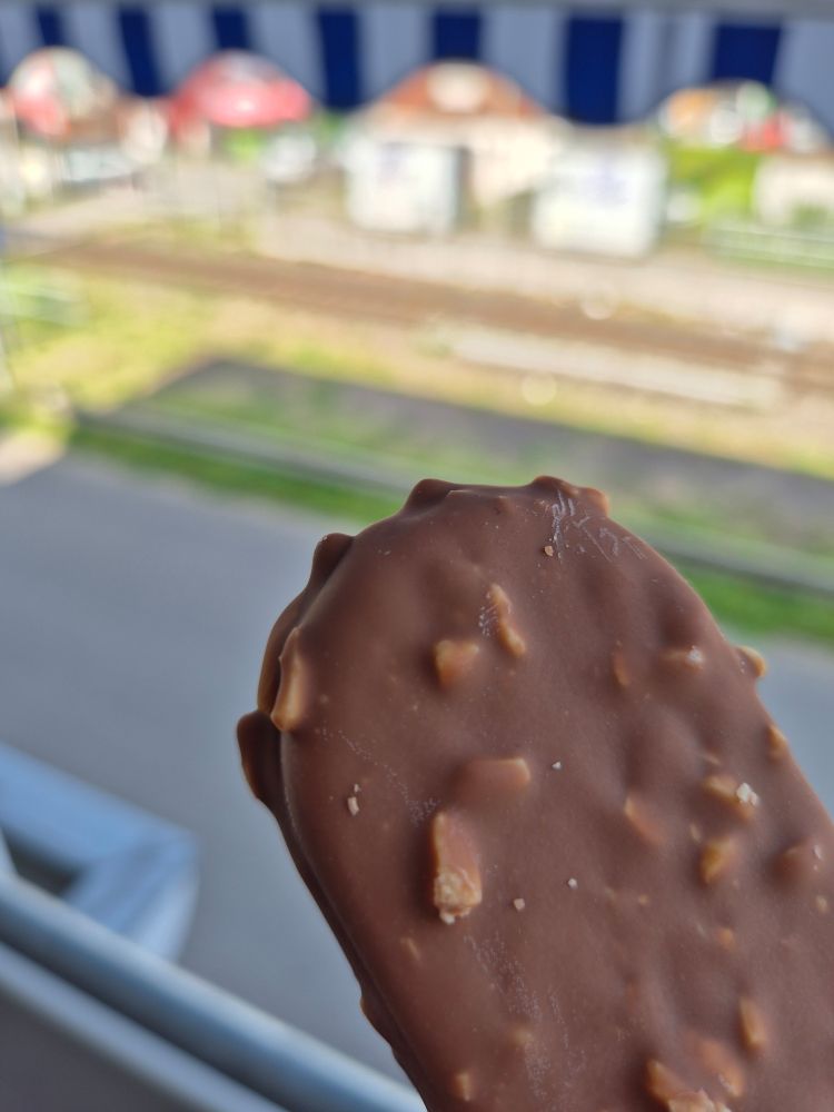 Ein Eis am Stil mit Schokoladenüberzug. Im Hintergrund unscharf der Blick von meinem Balkon 