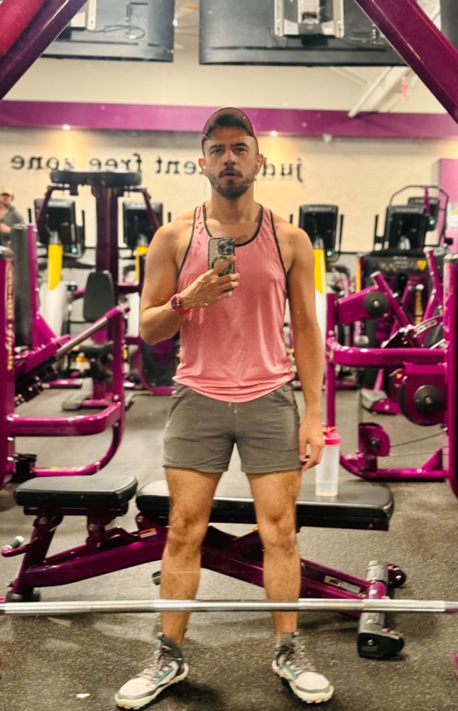 OP taking a mirror selfie at a gym standing in front of the smith machine, wearing pink tanktop, pink cap, and grey shorts.