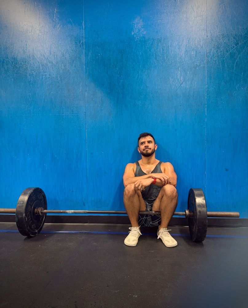 OP wearing green tank top and black/white shorts, sitting on a barbell with a plate on each side in front of a big blue gym wall. OP is looking to the side, smiling.