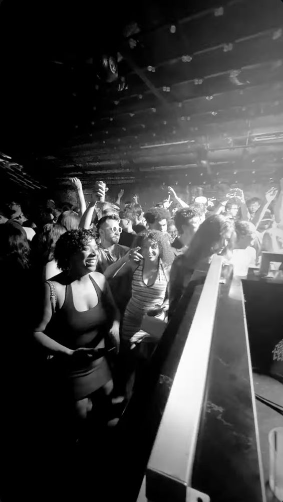 a photo of the crowd from the perspective of the DJ booth, people smiling, hands up, looking like they are having a fun time.