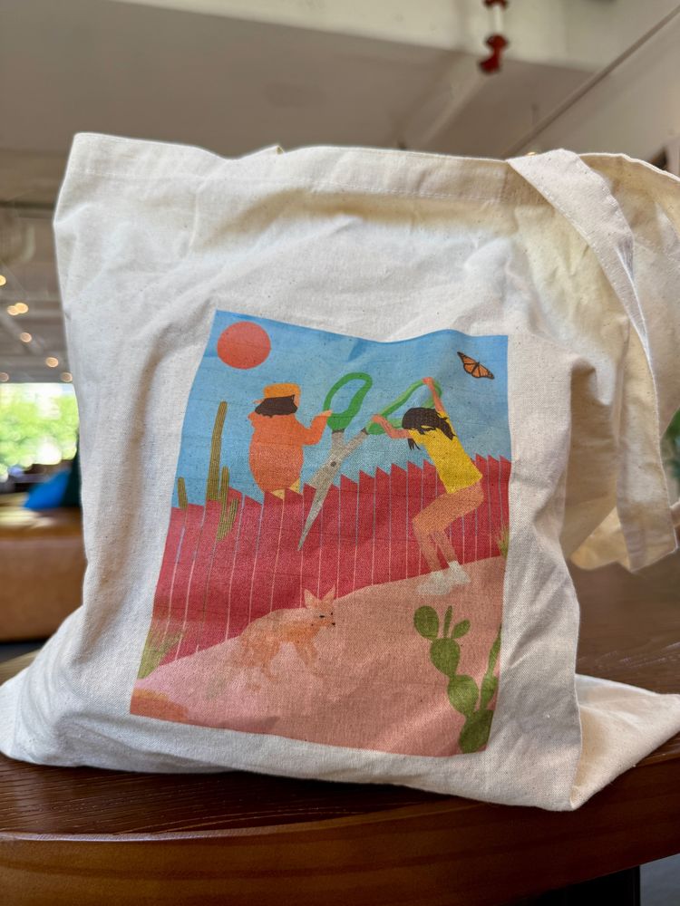 Photo of a tote bag with a drawing as its main visual. The drawing shows a red border fence being cut by two children sitting on the fence holding a giant pair of green scissors. Underneath them is a fox on the ground and part of a cactus. Above them is a red sun, a blue sky, and a monarch butterfly (a symbol used by immigrant rights activists)