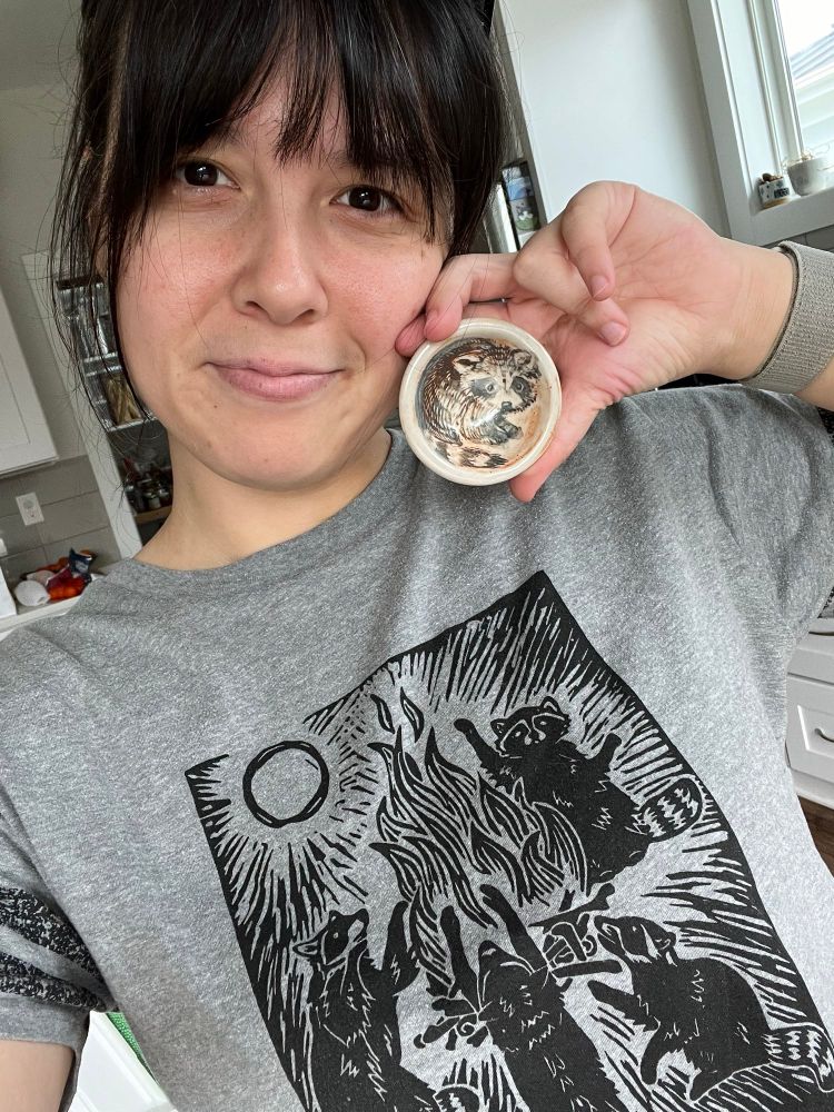 Brown haired and brown eyed artist is holding a small ramekin near their face with a baby racoon painted inside it, while wearing a grey shirt with raccoons around a campfire. The shirt is made by Critter Butts, artists in Charlottesville, VA. 