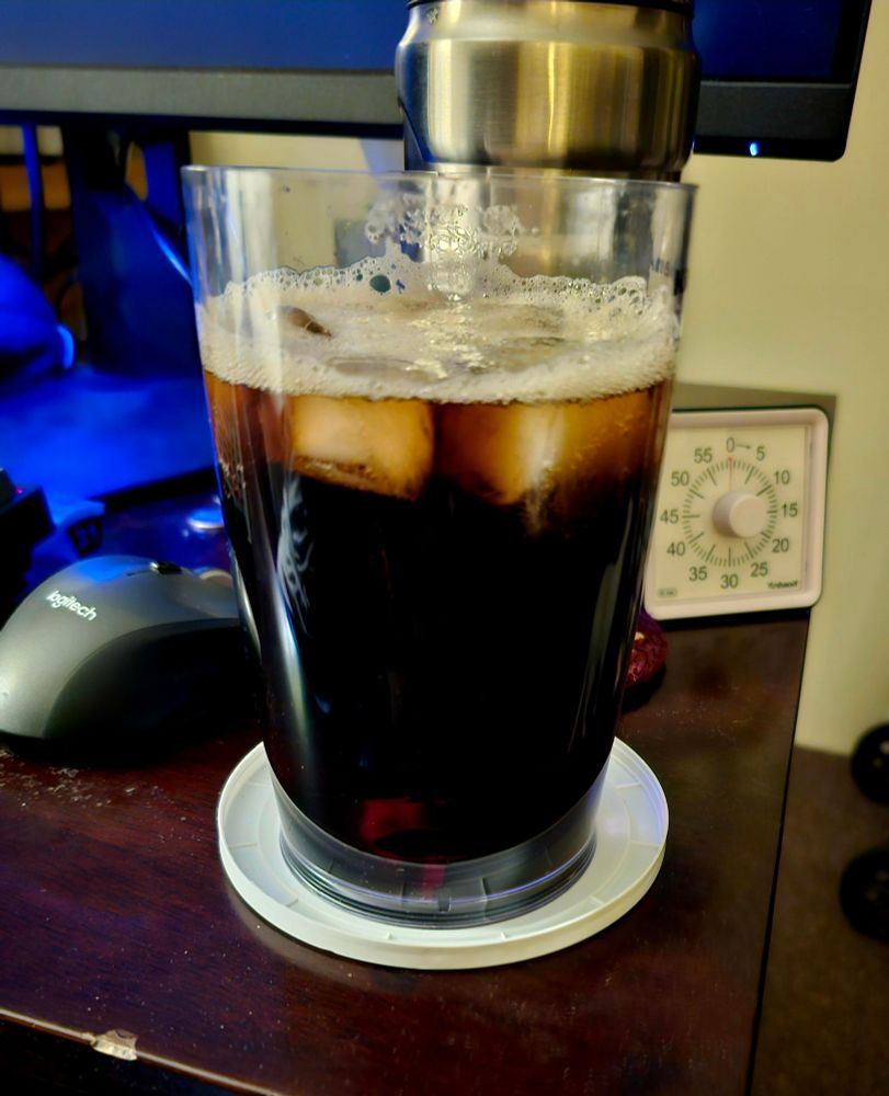 A tall glass filled with a Jack and Coke cocktail sits on a white coaster atop a wooden desk. The dark mixture shows distinct layers with ice cubes floating in it, creating a foamy head at the top. A metallic can is partially visible at the top of the frame. The scene is lit by blue ambient light from what appears to be a monitor, with a Logitech mouse visible to the left and a timer or clock device to the right of the glass.