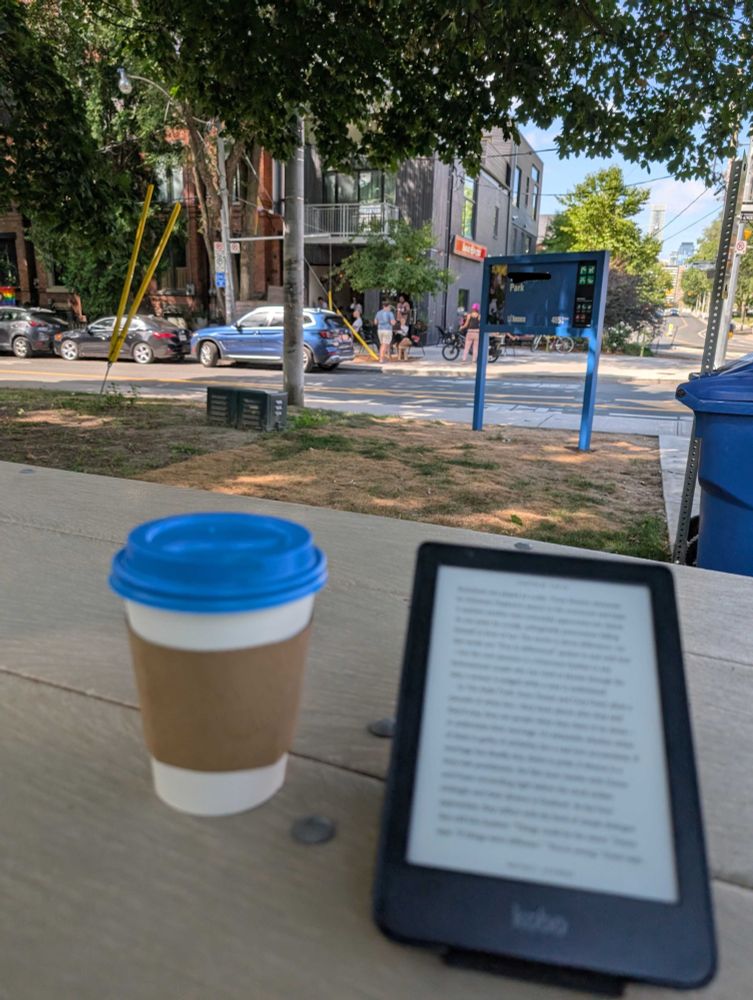 A coffee and an ereader on a picnic table in the foreground, trees and a lively coffee shop on the corner in the background