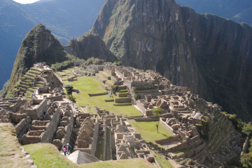 The remains of buildings made of rocks on a mountain with a huge cliff and mountains in the background
Through the middle is a grass pathway
A few tourists can be seen standing