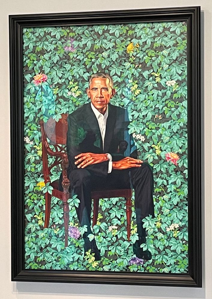President Barack Obama in a black suit - no tie, collar slightly open - is seated in a brown chair, sitting slightly forward with his arms crossed. He is surrounded by flowers.