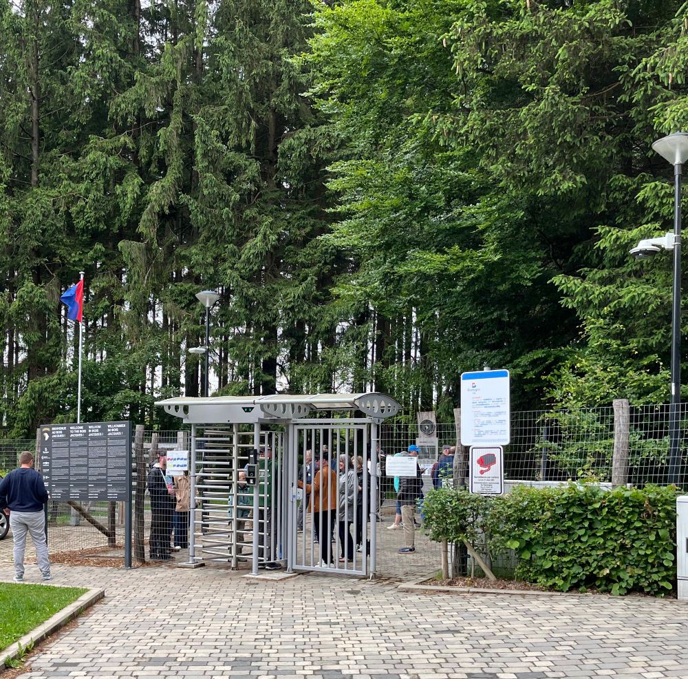 The entrance has a barred door to the right and a turnstile to the left with an entrance keypad hanging a bar in front. The wood are in the background with several tourist behind the entrance. A man reads an information board in four languages.