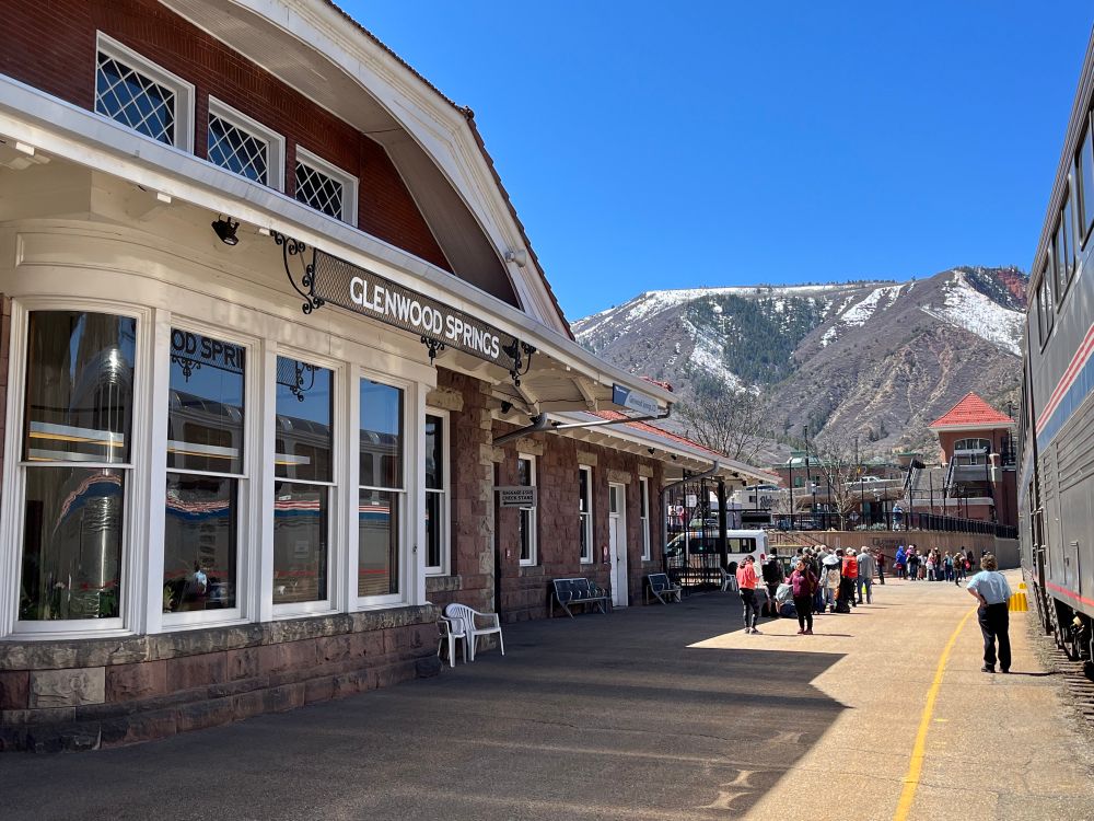 The train to the right can be seen in the reflection from the windows of the train station to the left. Glenwood Springs is displayed above the station. There are people standing outside and a mountain in the background.