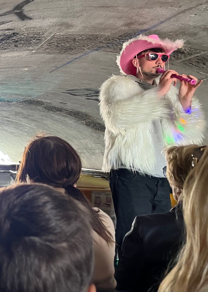 A tour guide standing at the front of the boat with the tourists sitting in the front seats
The guide is wearing black pants, white fluffy jacket with a pink cowboy hat, pink sunglasses. He is playing a pink flute.
The boat is going under a bridge.