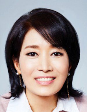 A professional headshot of Na Young Hee, a South Korean actress, with short, dark brown hair styled in a layered cut. She has a warm smile, well-groomed eyebrows, and natural-looking makeup. She is wearing dangling earrings and a white collared shirt with a soft pink blazer. The background is a plain, light gray color, and the lighting is even, highlighting her features clearly.