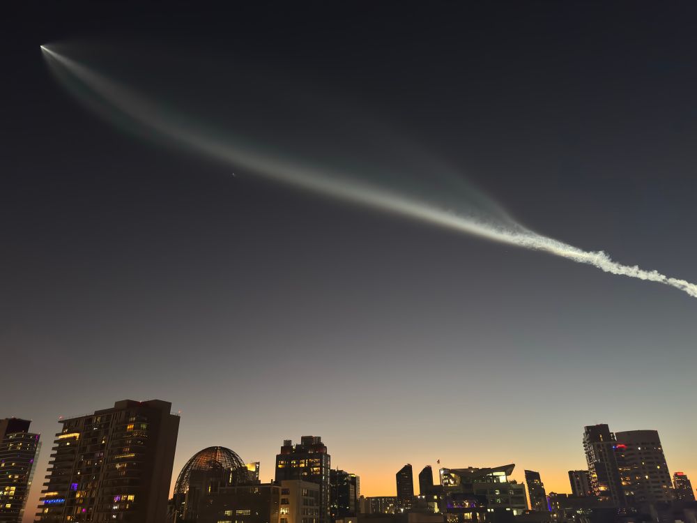 A SpaceX Falcon rocket jetting across the sky. We see the San Diego skyline. It is dusk. The trail of the rocket shine bright white, lit by the sun already tucked bellow the horizon. 