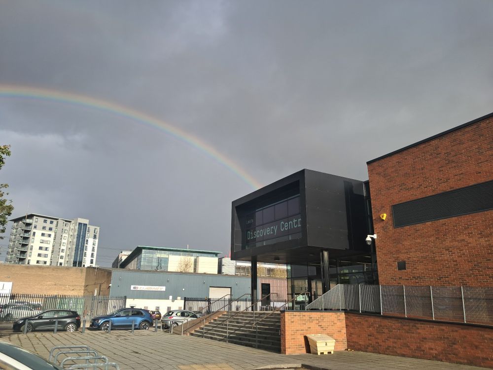 Large brick and black building on right, with road and warehouses to left and in front, grey skies and a rainbow.