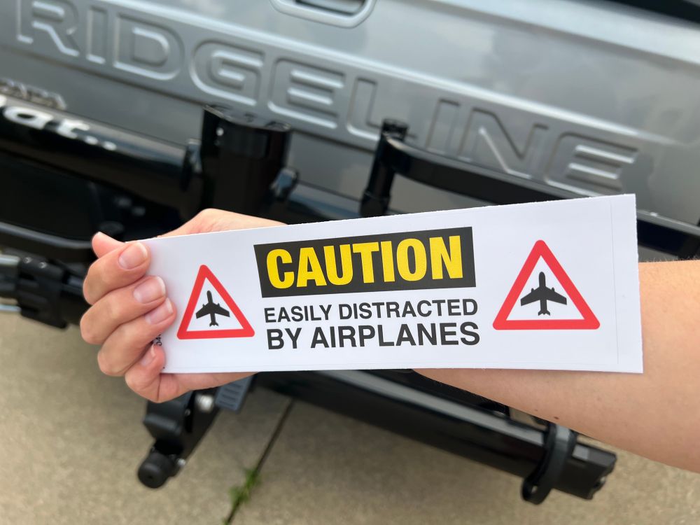 A bumper sticker that says “CAUTION easily distracted by airplanes”. It’s being held up in front of the back of a Ridgeline truck with a bike rack on it. It’s not my truck; it’s a rental.