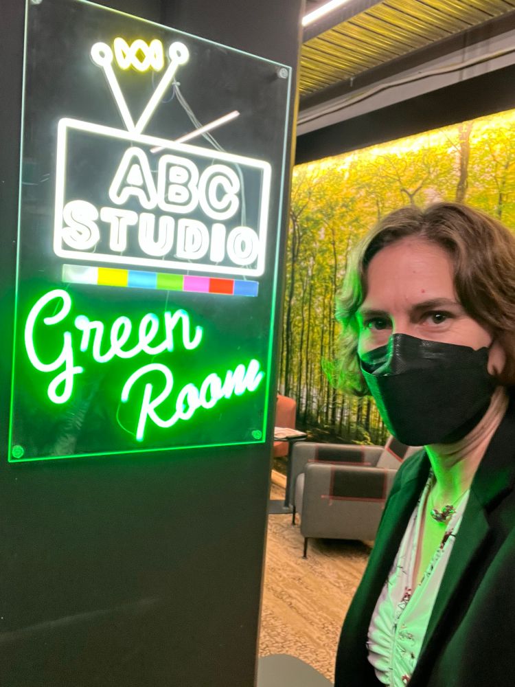 A selfie of me wearing a black mask standing next to a neon sign that says “ABC Studio Green Room”. Behind me are some chairs and a wall that is covered in wallpaper of a forest 