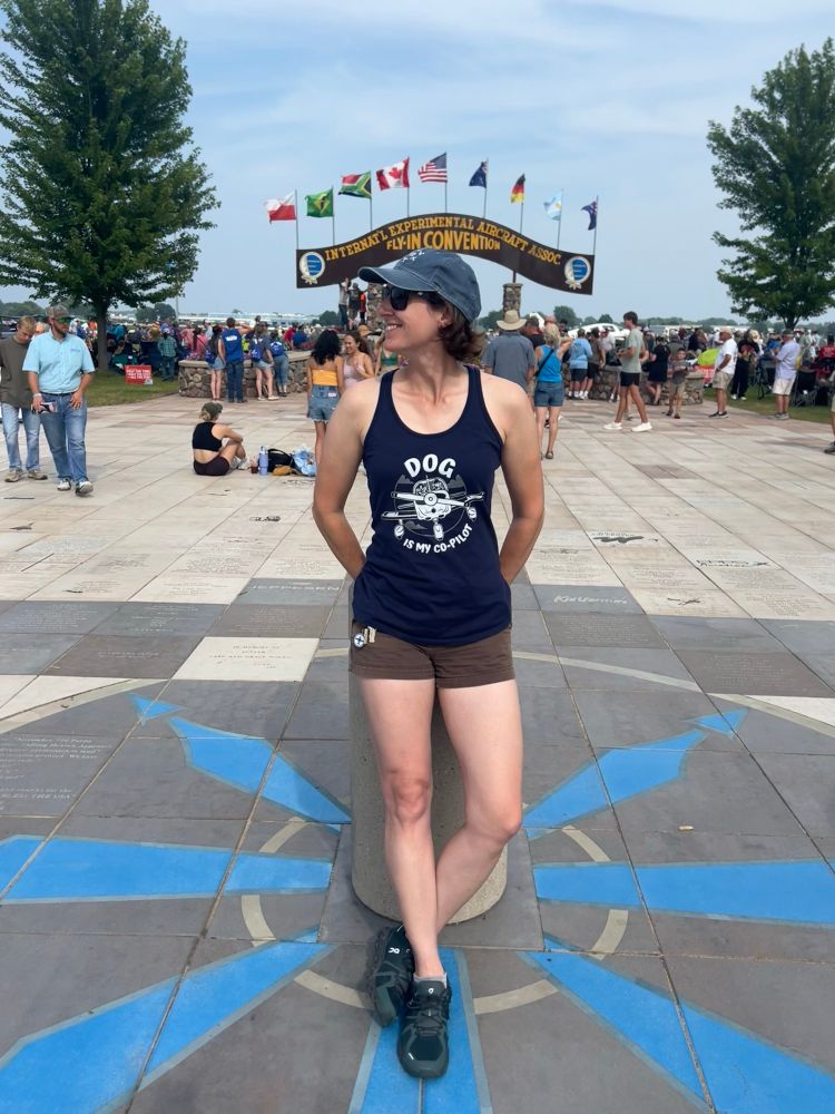 Another view of me standing in front of the AirVenture arch, this time a longer view so you can see the full length of the tank top. I’m wearing shorts and looking to the side.