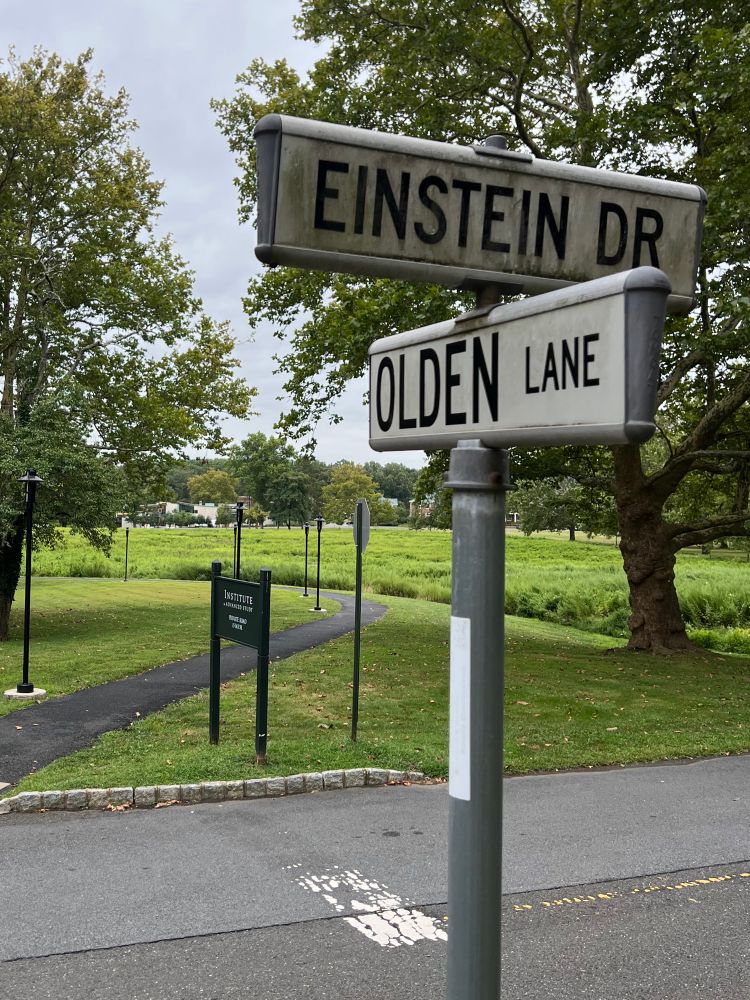 The road signs for Einstein Drive and Olden Lane, with the Institute in the background surrounded by trees 