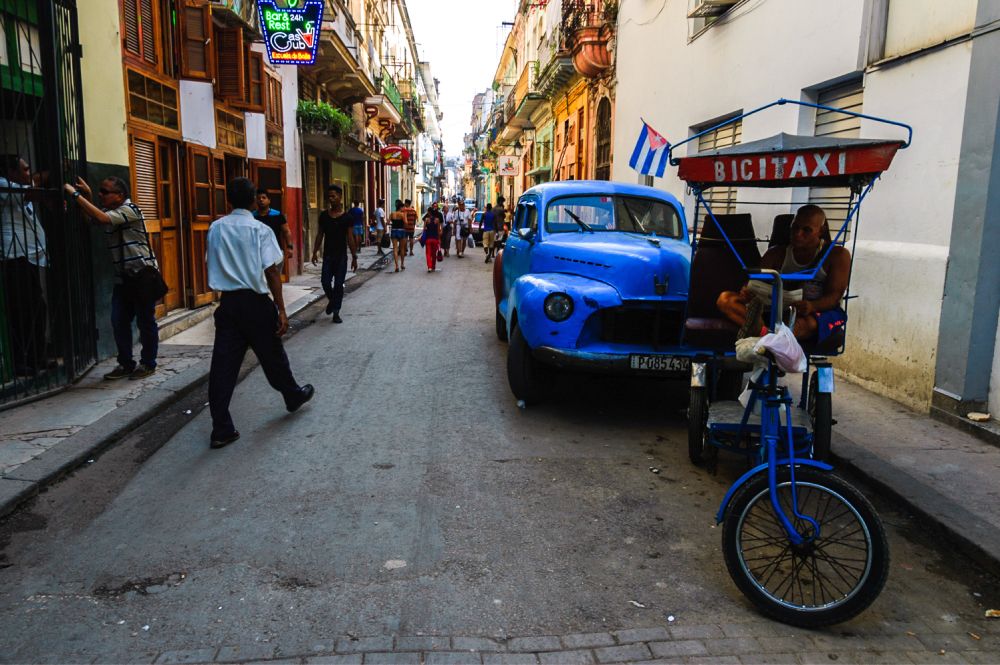 A lively street scene in a historic district with colorful old buildings. A blue vintage car is parked beside a bicycle taxi (bicitaxi) with a Cuban flag. People walk along the street, and a neon sign for a bar is visible on the left.
