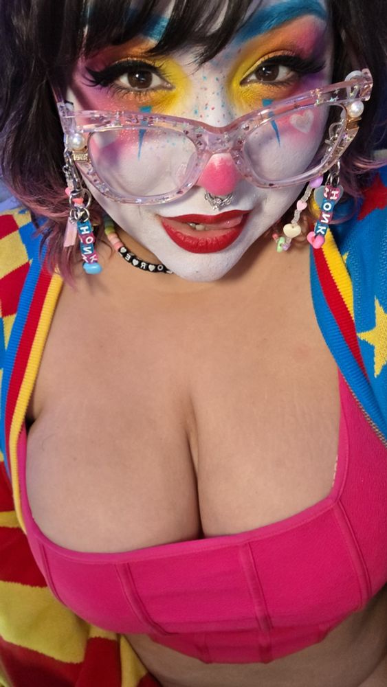 Missy in clown makeup and glasses. She has a low cut shirt on and a cardigan. She is smiling. 