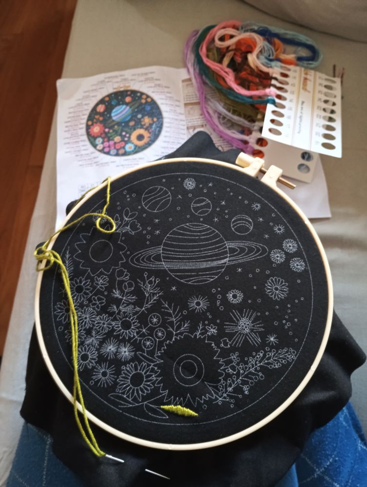 Tela negra con un dibujo de planetas y flores preparado para bordar, con una hoja bordada en verde en la parte inferior.