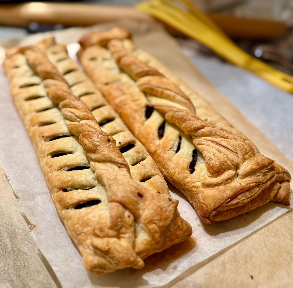 photo of two mushroom chestnut wellingtons--puff pastry folded around a mushroom walnut chestnut filling (one of them) and a mushroom parsnip chestnut filling (other one of them for someone who requested no nuts). simply folded pastry is decorated with a braid down the center and cut slits to let the steam out.  ngl house smells v good. 