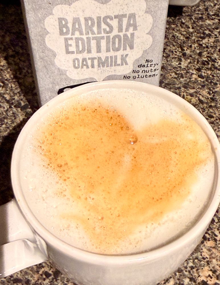 A photo of a cappuccino in a white mug with oatly barista edition oat milk in the background.