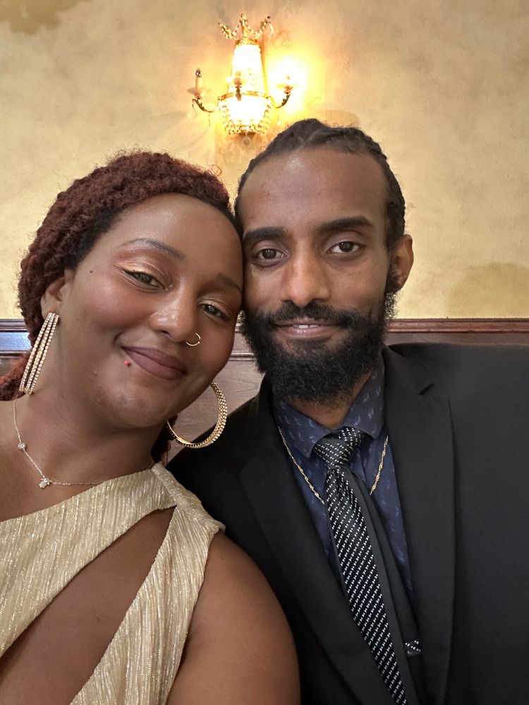 A selfie of me and my husband. I am on the left in gold, he is in a suit and tie with his locs braided back (looks like Nipsey Hussle)