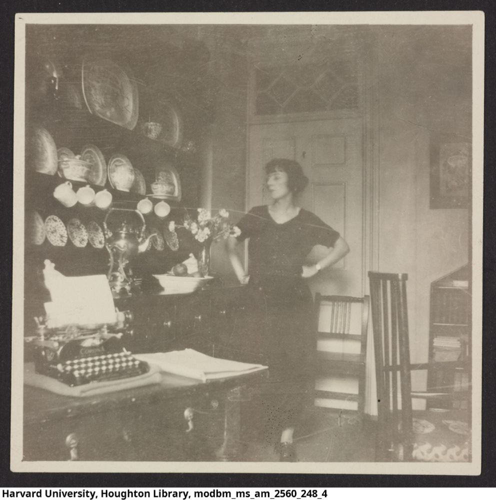 black and white photograph of a woman (white, dark hair with bangs pulled back into a bun in a long dark dress and dark shoes) stands with her hands on her hips behind a desk with a corona typewriter and next to a beautiful hutch holding displaying fine china, a silver samova, and a vase of flowers