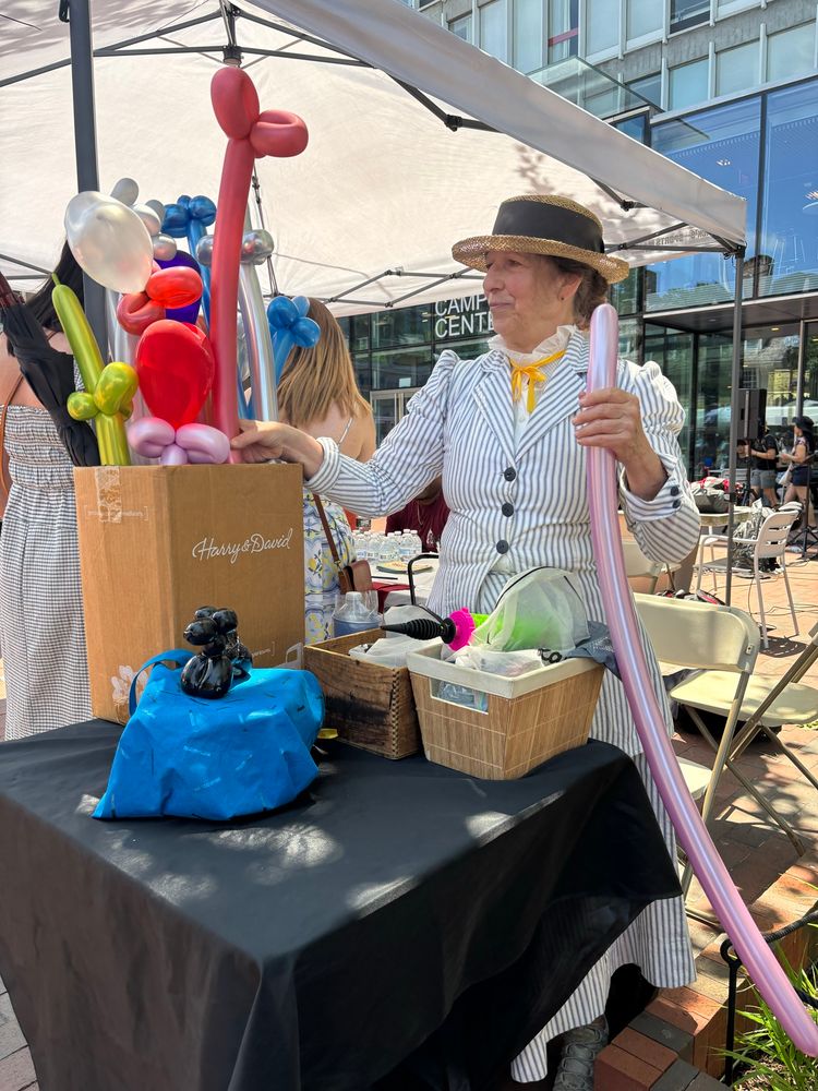 A woman in a pinstripe jacket and skirt and Panama hat holds a large pink balloon under a white canopy 