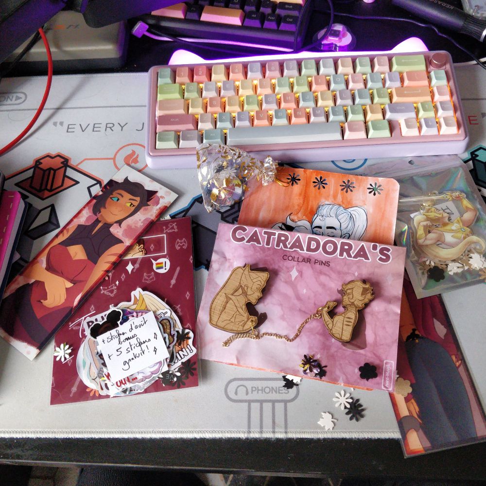 Un bureau en bazar sur lequel est étalé le contenu de ma commande à Aure : 
2 marques pages Catra
2 broches de col Catra et She Ra en bois
Un pack de stickers
Un badge Nimona non binaire
Un porte clé She Ra