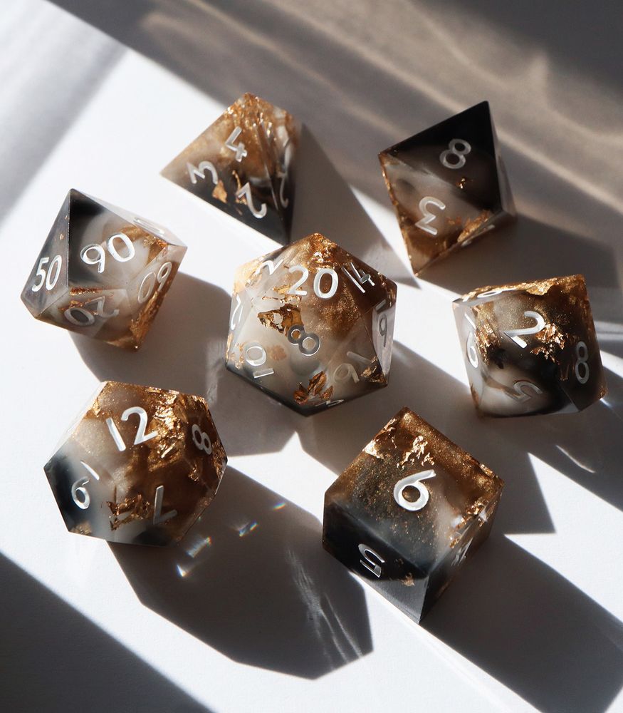 Full set of Allured arranged in a circle with the D20 in the center. The dice are a marbled mix of white and black resin with sparkling gold glitter swirled around and gold leaf throughout. The numbers on the dice are white.