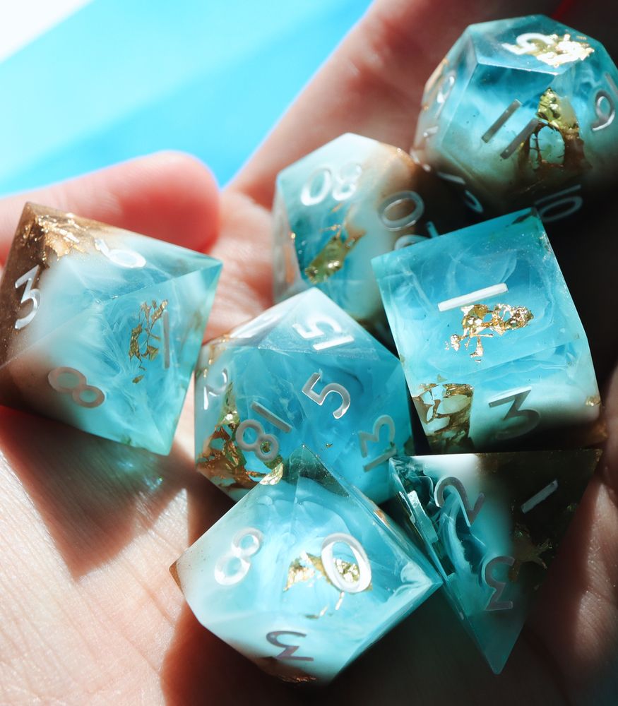 Full set of Riptide held in a person’s hand with the sunlight catching the dice. All the dice are a mix of cerulean blue, white, and tan resin to mimic a sandy shore. Ripples of white resin smoke resemble waves. There are gold foil inclusions.