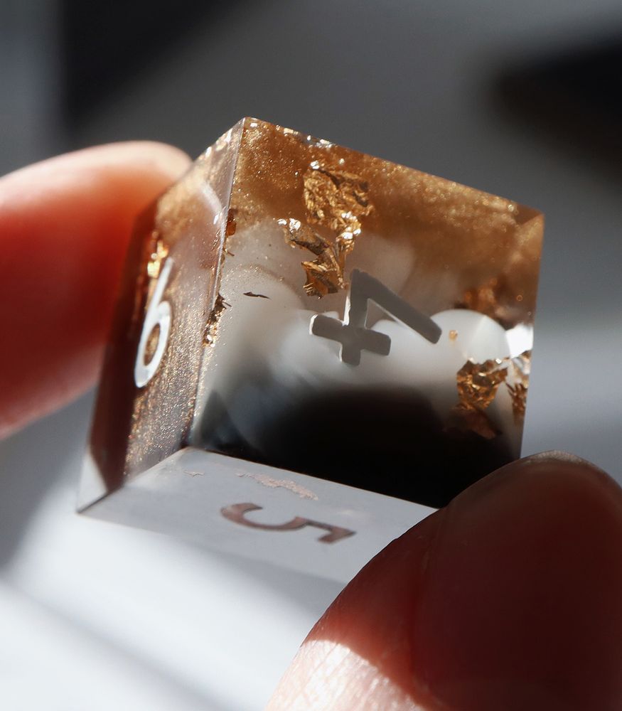 Closeup of the Allured D6 held in between two fingers. The die is half clear resin half a smokey mix of black and sparkling white resin. There are flakes of gold leaf and glittering gold throughout. The numbers on the die are white.