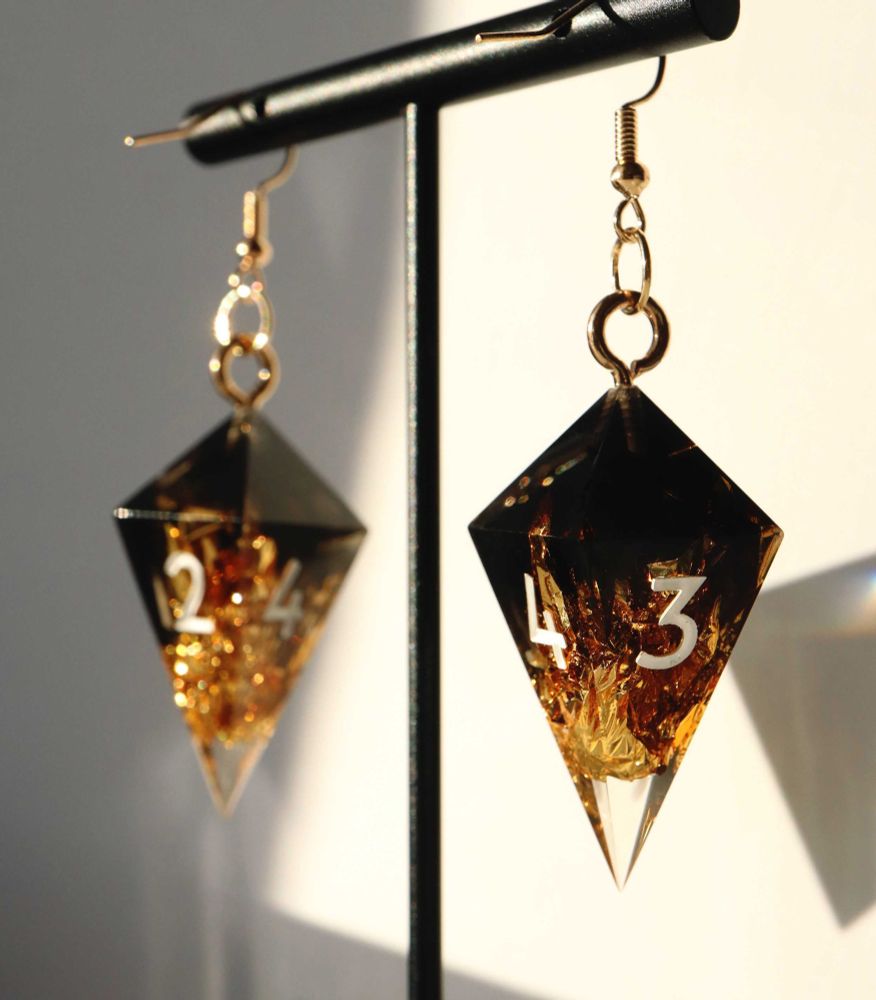 Brutus D4 Earrings handing on an earring stand. The pendants are a crystal D4 shape of black and clear resin with gold foil throughout, making a gradient from black to clear. The numbers on the dice are white.