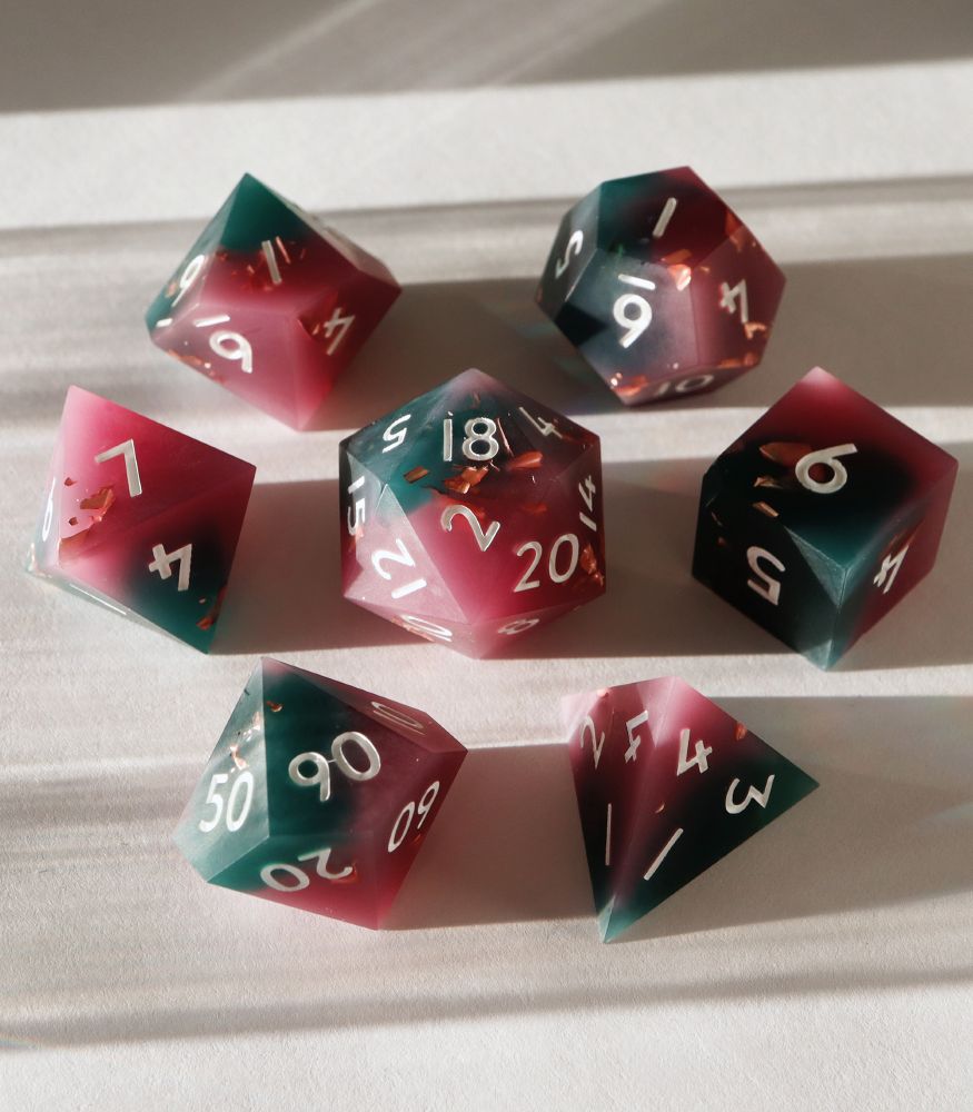 Full set of Disgraced 7 piece dice set, arranged in a circle with the D20 in the center. All the dice are a half and half mixture of pink and teal matte resin with small inclusions of copper foil. The numbers on the dice are white.