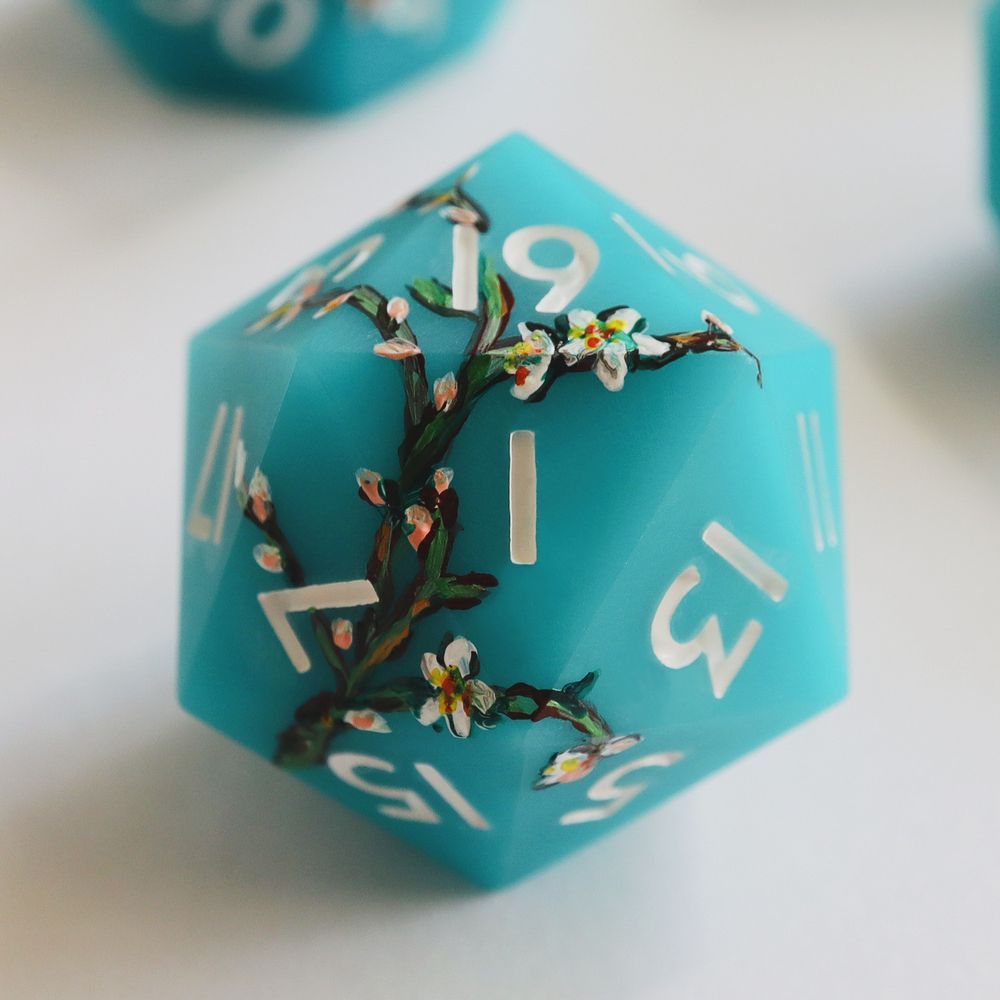Single light blue matte d20 on a white background. The die has impressionist-style illustrations of white blossoms and green and brown branches painted around it. The numbers on the die are white.