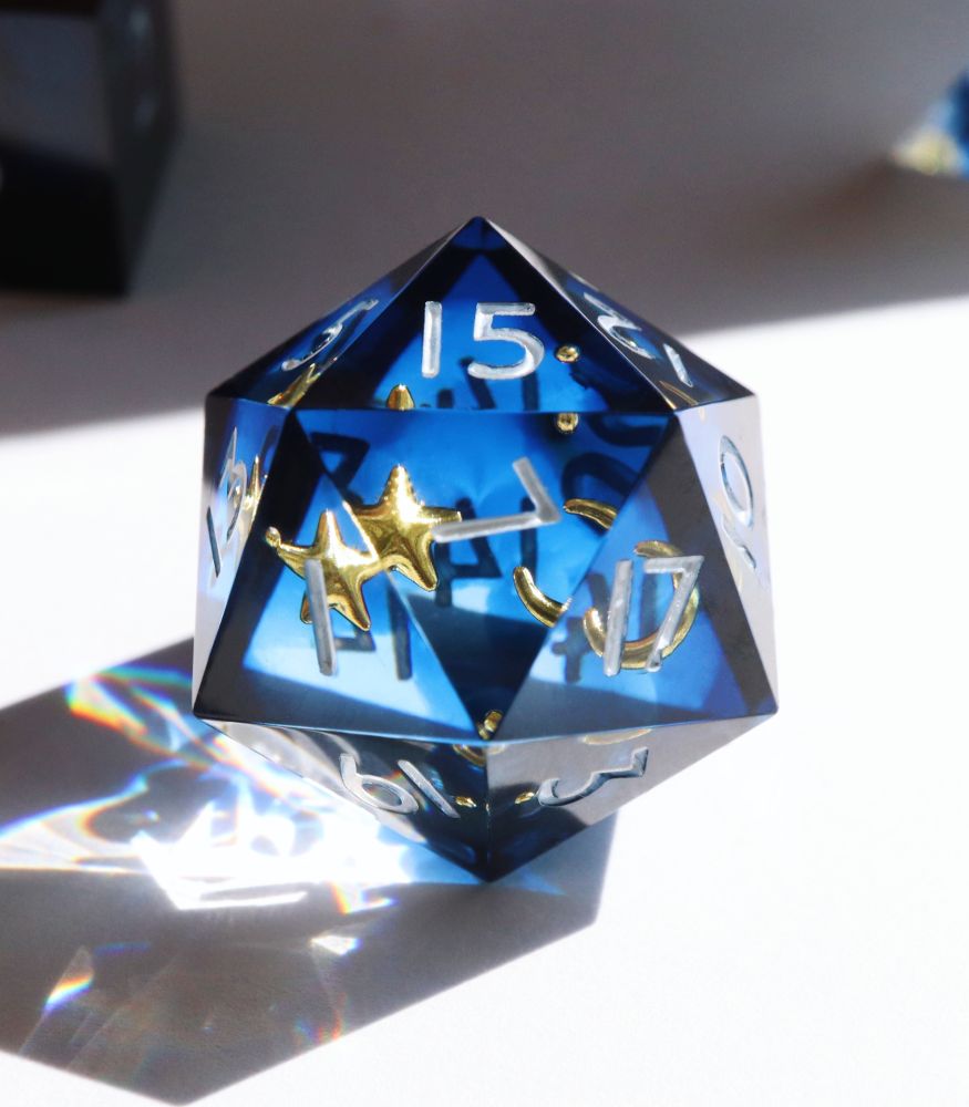 Closeup of the Etoile Royale D20. The die is a mix of clear resin and transparent dark blue resin with inclusions of gold moons and stars. The numbers on the die are white.
